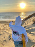 Un homme avec un sweat à capuche blanc porte une planche de surf